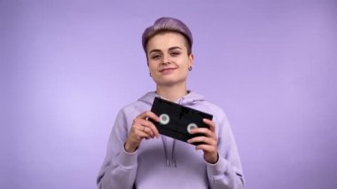 Neşeli, genç, yetişkin, modaya uygun, modaya uygun, elinde klasik VHS kaset tutan, elinde manyetik kaset olan, kapalı alanda mor arka planda izole edilmiş gülümseyen kameraya bakan bir kadın nesli. Sentetik dalga