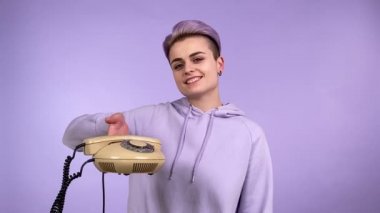 Boyalı kısa saçlı, menekşe kapüşonlu, elinde klasik kablosuz telefon, çevirme numarası olan, izole edilmiş mor arka planda gülümseyen modern yetişkin bir kadın. Çevirmeli telefon kullanan büyüleyici lgbt kişisi
