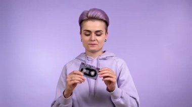 Modaya uygun boyalı kısa saçlı neşeli genç bir kadın elinde klasik bir kaset tutarak, zevkle manyetik bandı çıkarıyor, mor arka planda izole edilmiş stresi hafifletiyor. Sentetik dalga konsepti