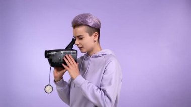 Mor renkli saçı olan genç bir insan, eski VHS video kamerasını kullanıyor, görüntü bulucuya bakıyor, kapalı alanda mor arka planda izole edilmiş klasik kamera ile çekim yapıyor. Sentetik dalga konsepti