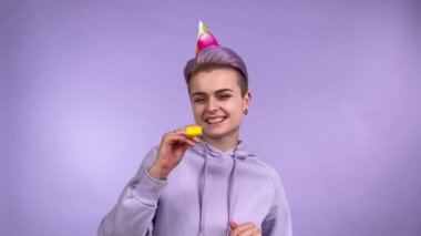 Neşeli, kısa boyalı saçları olan, parti şapkası takan, parti kornası çalan, doğum günü partisi malzemelerini kullanan, olumlu duygularını paylaşan genç bir kadın. Mor üzerine izole edilmiş kapalı stüdyo çekimi