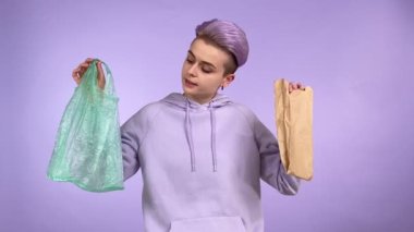 Mod genç yetişkin kadın nesli Z, kısa boyalı saçlı plastik ve kağıt torba tutuyor, paketleri karşılaştırıyor, çevre dostu geri dönüşümlü ambalajı mor arka planda izole ediyor.