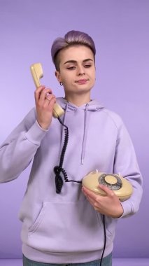 Mor kapüşonlu bir kapüşonlu giyen, elinde klasik şifreli bir telefon olan, kapalı alanda izole edilmiş, çevirmeli telefon kullanan genç bir kadının dikey görüntüsü.