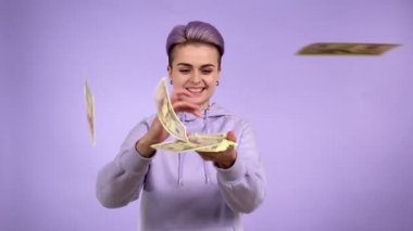 Mutlu genç insan geneli Z trendi bir grup parayı doğrudan kameraya atıyor, banknotları dağıtıyor, el kaldırıyor, şans piyangosuna seviniyor, mor arka planda izole edilmiş para karıyla övünüyor.