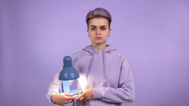 Yüzünde boş bir ifade olan genç bir kadın eski moda masa lambasını açıp kameraya bakıyor. Mor arka planda izole edilmiş menekşe kapüşonlu kısa saçlı bir kadın.