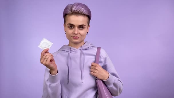 短い染めの髪を保持コンドームを示す正の若い大人の女性は 屋内で紫色の背景に隔離されたカメラを見て ハンドバッグに避妊機器を入れて モノクロームの概念 — ストック動画