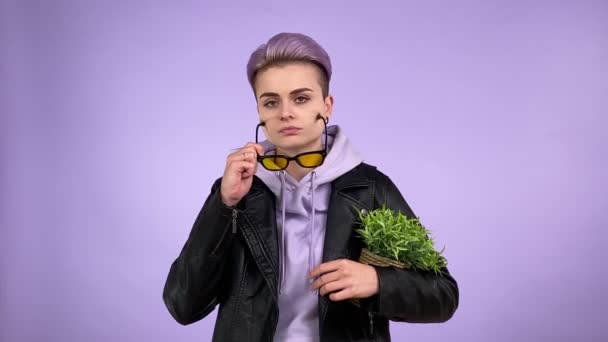酷酷的女同性恋 穿着时髦的皮夹克 拿着绿色盆栽 带着干巴巴的愤世嫉俗的眼神看着相机 戴着隔离在室内紫色背景的太阳镜 — 图库视频影像