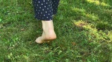 Rear view of female legs strolling along green lawn at sunrise outdoor. Barefoot woman enjoying morning freshness by walking grass at garden. Relaxation, healthy lifestyle concept