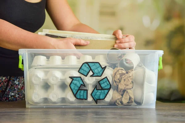 Female hand sorting waste paper. Transparent plastic container with recycle sign full of wastepaper on wooden table on blurred background. Recycling old paper products reused. Environment conservation