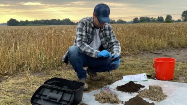 Agronomy scientist conducting soil research by using digital measuring device, entering data into tablet at agricultural grain field in rays of sunrise. Environment protection, soil certification