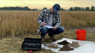 Male agronomist in squatting position taking notes in sampling information sheet, glancing at heaps of different soil types background of grain field at dawn. Environment research, soil certification
