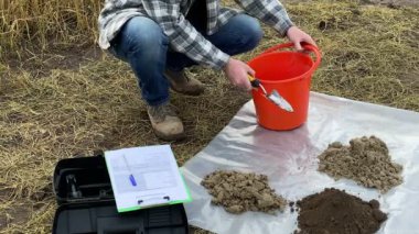 Male agronomist taking soil sample out from bucket with garden trowel on plastic underlay outdoors. Heaps of different soil types arranged on test area. Environment research, soil certification