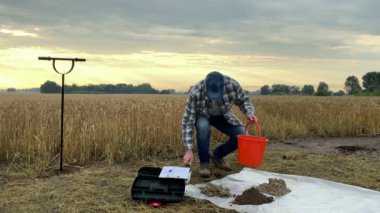 Male agronomy specialist pouring soil sample from bucket to underlay, taking notes in sampling information sheet outdoors. Scientist preparing for testing at field. Environment research, certification