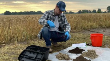 Agronomist preparing for soil measurements with digital device, sticking tester probe into sample, measuring pH temperature moisture of ground at agricultural grain field at warm summer dawn.