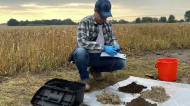 Agronomy scientist conducting soil research by using digital measuring device, taking notes in sampling information sheet at agricultural grain field in rays of sunrise. Soil certification concept