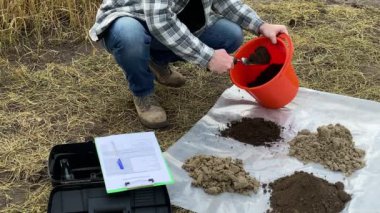 High angle view of agronomist taking out soil sample from bucket, taking notes in sampling information sheet outdoors. Male scientist preparing for testing at field. Environment research certification