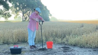 Female agronomy specialist soil sampling by probe sampler putting material in bucket at agricultural grain field dawn. Woman agriculturist working outdoors. Environment research, soil certification