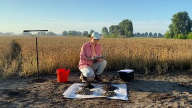 Woman agriculturist soil sampling, taking notes in digital tablet at agricultural grain field at sunrise. Female agronomy specialist working outdoors. Environment research, soil certification
