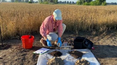 Female scientist testing soil sample outdoors, using laboratory equipment, performing soil certification at agricultural grain field sunrise. Woman agronomist working at grounds. Environment research