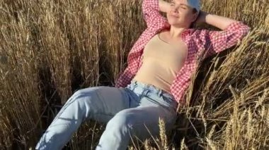 Carefree young adult woman relaxing lying back at wheat field at sunrise. Beautiful female farmer expressing wellbeing, happiness, positive emotions outdoors. Countryside lifestyle. Moving shot