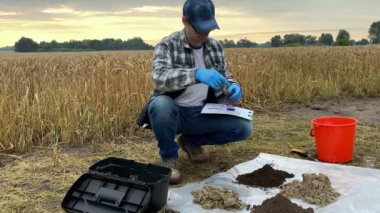 Male agronomist preparing for measurement of soil samples by using digital soil tester outdoors, holding sample information sheet. Professional farmer working at agricultural field at sunrise.