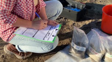 Closeup of female agronomist preparing soil samples for laboratory analysis, writing in information sheet outdoors at sunrise. Woman farmer taking notes in form, working at field. Soil certification