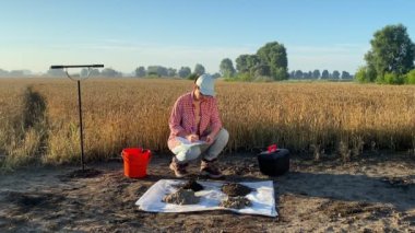 Female farmer preparing soil sample for laboratory analysis, taking notes information sheet at sunrise outdoors. Woman specialist performing environment research by sampling soil at agricultural field