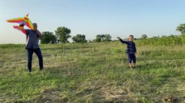 Positive father spending time with son by flying kite at meadow sunset. Little boy running with kite, enjoying playtime with parent outdoors. Happiness in simple living