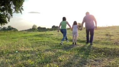 Silhouette of family with one child walking into sunset at meadow. Happy family spending time together outdoors. Little girl going near parents hand in hand, jumping in gold lights of summer sun