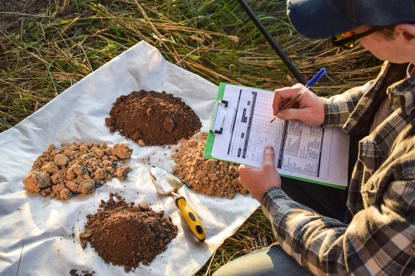 High angle view male agronomist preparing soil samples for laboratory analysis, writing in information sheet outdoors at sunrise. Man farmer taking notes in form, working at field. Soil certification