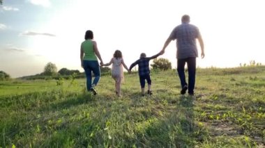 İki çocuğun birlikte dışarıda vakit geçirdiği bir aile manzarası. Baba, anne el ele tutuşuyor oğul kız gün batımına doğru yürüyor çayır üzerinde. Mutlu ebeveynler doğadaki çocuklarla vakit geçiriyorlar.