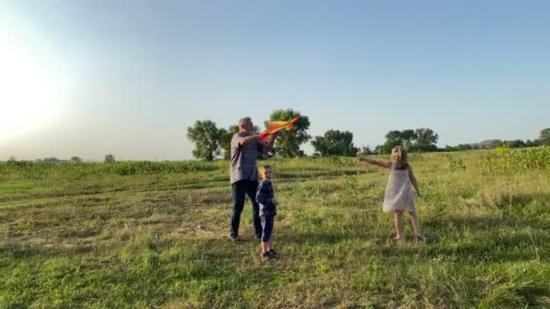 有两个孩子的家庭在自然日落时度过闲暇时光 爷爷教孩子们在户外放风筝 小女孩带着玩具在乡间路上跑 简单生活中的快乐 — 图库视频影像