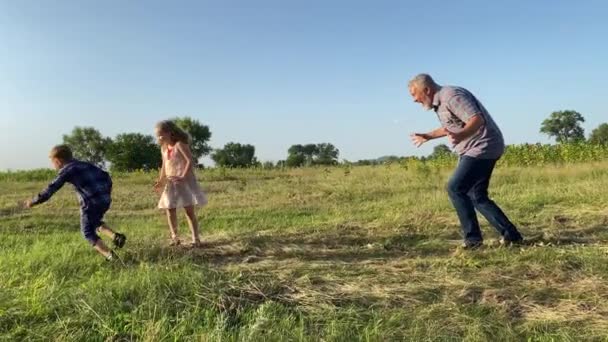 屋外で遊んでいる2人の子供を持つ家族 田舎で娘と息子と遊んでいる陽気な父親 祖父は日没時に子供たちと余暇を過ごしています 簡単な生活の中で幸せ — ストック動画