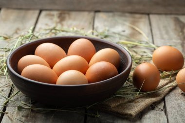 Ahşap arka planda kilden bir kasede tavuk yumurtası, saman etrafa saçılmış. Sağlıklı gıda konsepti. Çiftlik işçiliği ve bakımı.