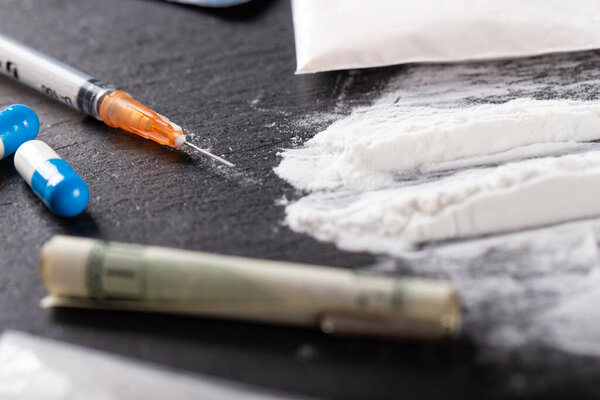 Cocaine or other illegal drugs, white powder, money, pills, syringe on black background. Selective focus.