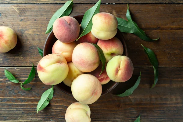 Ahşap bir kırsal masada, kil bir kasede taze şeftali hasadı. Şeftali yaprakları kompozisyonda mevcuttur. Organik meyve, vejetaryen yemeği..