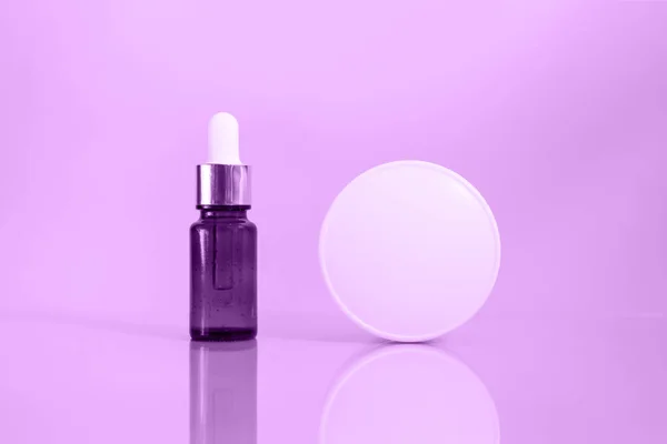 A glass dropper bottle with a pippette with white rubber tip and white jar of facial or body cream on the purple background. Nature Skin concept. Organic Spa Cosmetics. Close up.