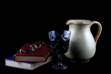 Şarap sürahisi, bir bardak şarap, eski kitaplar ve siyah bir arka planda gözlük.