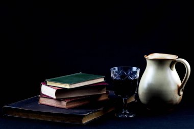 Old books with glass of red wine and earthenware wine jug on a black felt table cover