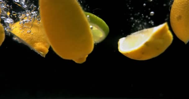 In Scheiben geschnittene Zitrone und Limette, die auf schwarzem Hintergrund ins Wasser fallen — Stockvideo