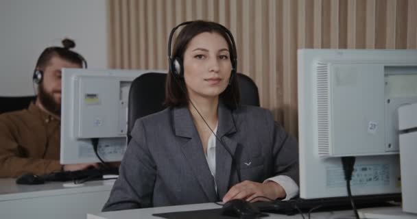Empleado del centro de llamadas - una mujer joven con auriculares sonriendo mira a la cámara — Vídeos de Stock