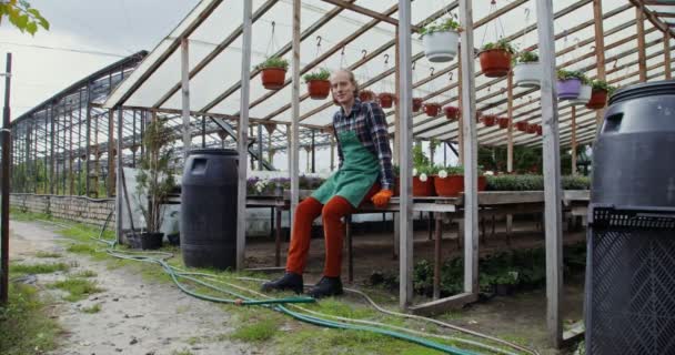 Un florista masculino se sienta en el borde de una mesa entre macetas de flores — Vídeo de stock