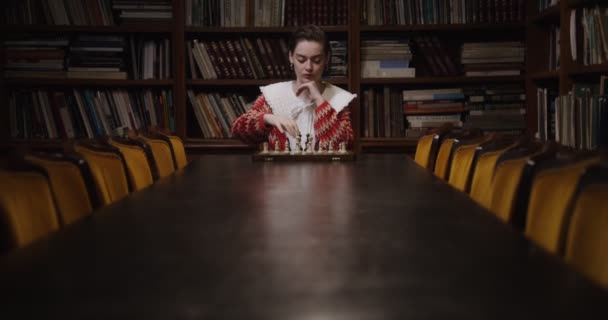 Mladá žena hraje šachovou partii sama, sedí u dlouhého stolu v knihovně — Stock video