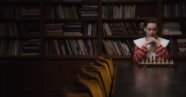 Młoda kobieta gra w szachy sama, siedzi przy długim stole w bibliotece — Wideo stockowe