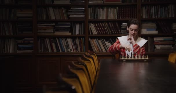 Mladá žena hraje šachovou partii sama, sedí u dlouhého stolu v knihovně — Stock video