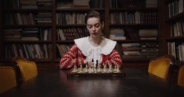 Een jonge vrouw speelt een schaakspel alleen, zittend aan een lange tafel in de bibliotheek — Stockvideo