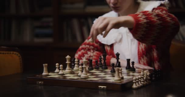 Una joven juega una partida de ajedrez sola, sentada en una mesa larga en la biblioteca — Vídeos de Stock
