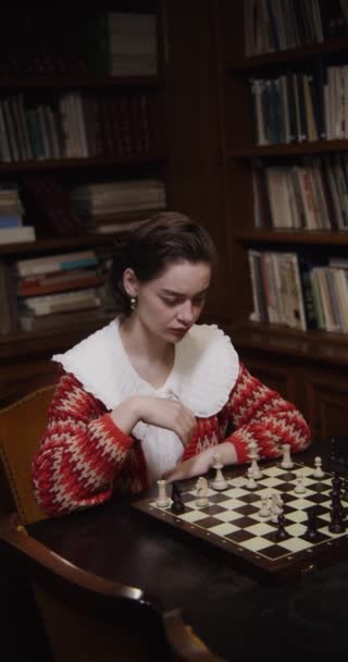 Μια νεαρή γυναίκα παίζει σκάκι μόνη της, καθισμένη σε ένα μεγάλο τραπέζι στη βιβλιοθήκη. — Αρχείο Βίντεο