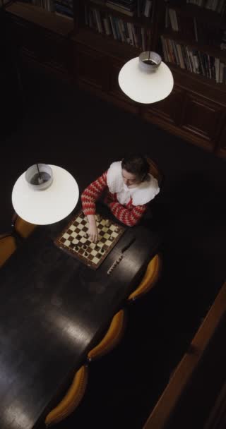 Una giovane donna gioca una partita a scacchi da sola, seduta a un lungo tavolo in biblioteca — Video Stock