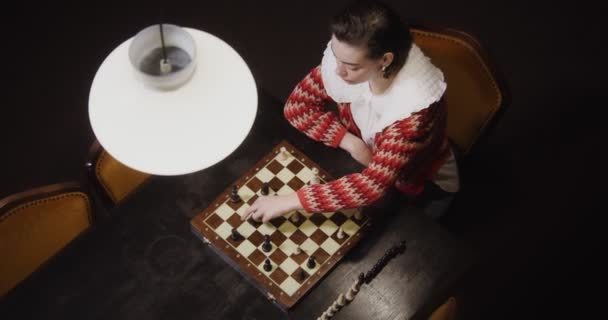 Una joven juega una partida de ajedrez sola, sentada en una mesa larga en la biblioteca — Vídeo de stock